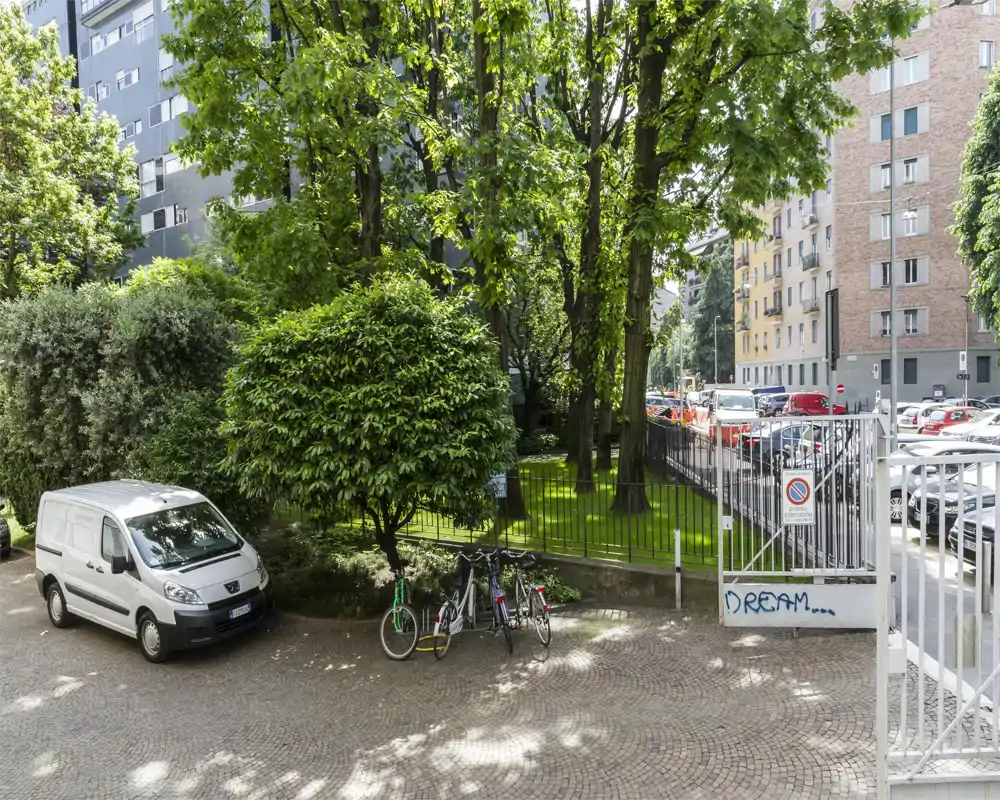 Patio/área de aparcamiento soleada con árboles, una furgoneta aparcada y varias bicicletas junto a una entrada con reja; agradable zona verde junto a edificios urbanos.