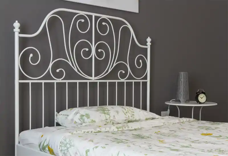 Dormitorio luminoso y ordenado con cabecero de metal blanco decorativo, ropa de cama floral y mesita con lámpara y despertador — perfecto para un anuncio acogedor.