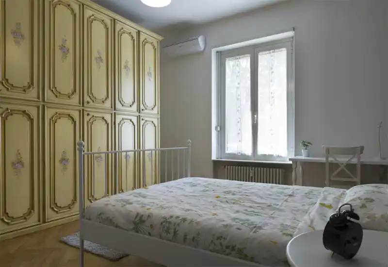 Dormitorio luminoso y bien compuesto con un armario vintage decorado, cama de hierro y ropa de cama floral; la luz natural de la ventana aporta una atmósfera tranquila y acogedora.