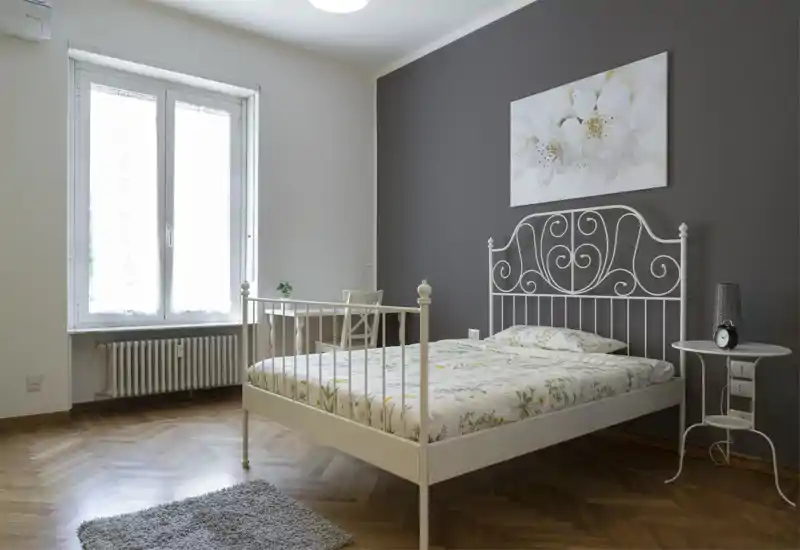 Dormitorio luminoso y minimalista con cama de hierro blanco, ropa de cama floral y pared oscura de acento; gran ventana que aporta luz natural.