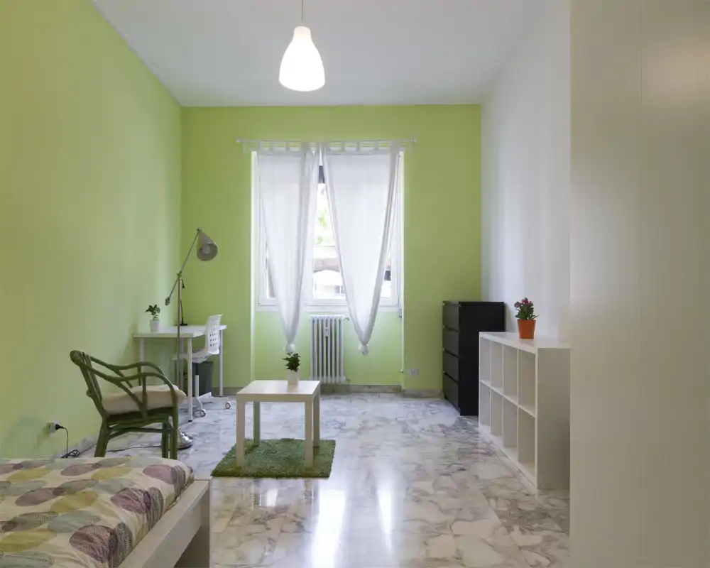Camera da letto luminosa e moderna con parete verde pastello, pavimento in marmo e mobili bianchi essenziali. Include un letto in vista nell'angolo, una scrivania, una poltrona e scaffalature, perfetta per una persona.