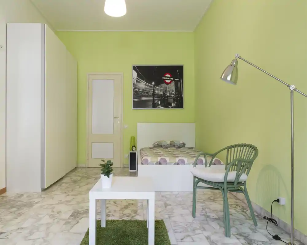 Camera da letto luminosa e moderna con letto matrimoniale contro una parete verde pistacchio, armadio bianco, comodino e una poltrona — pavimento in marmo e arredo semplice e fresco.