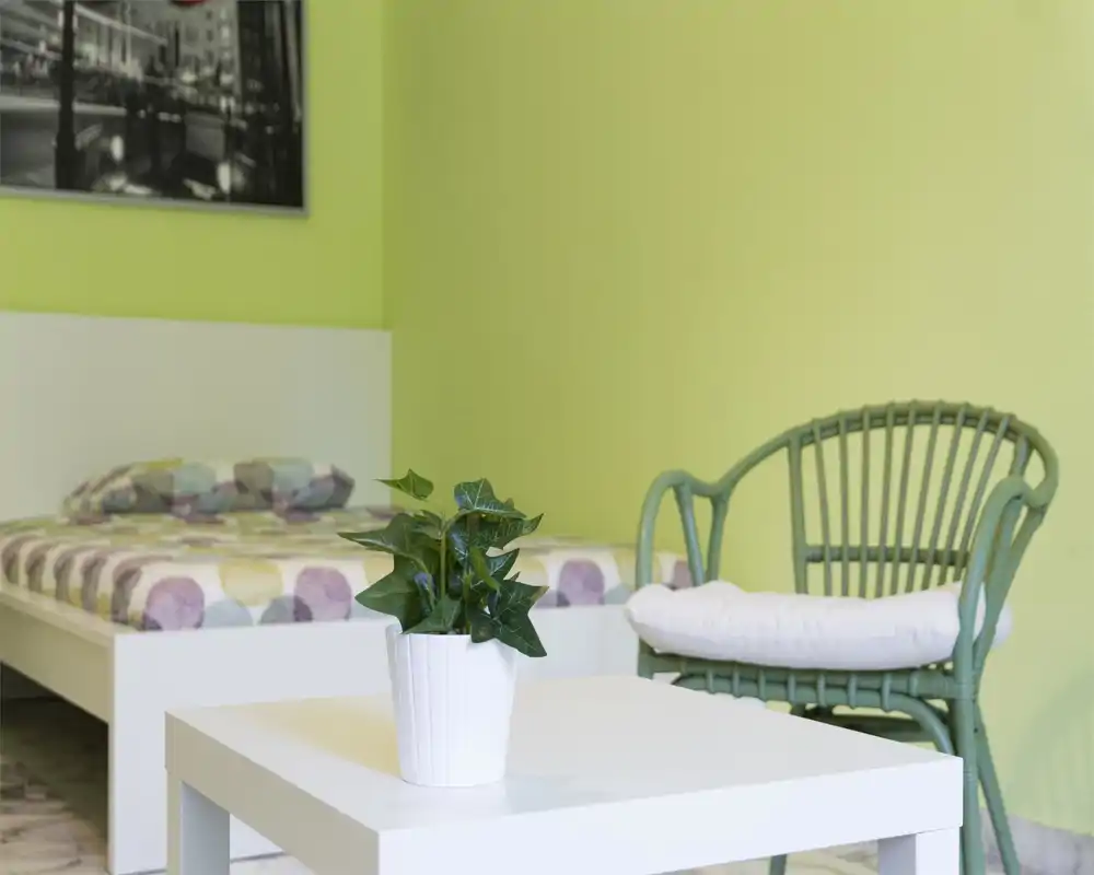 Camera da letto luminosa e tranquilla con letto bianco e biancheria a fantasia, piccolo tavolino bianco con pianta in vaso in primo piano e poltrona in midollino verde sul lato — arredamento fresco e minimal ideale per il relax.