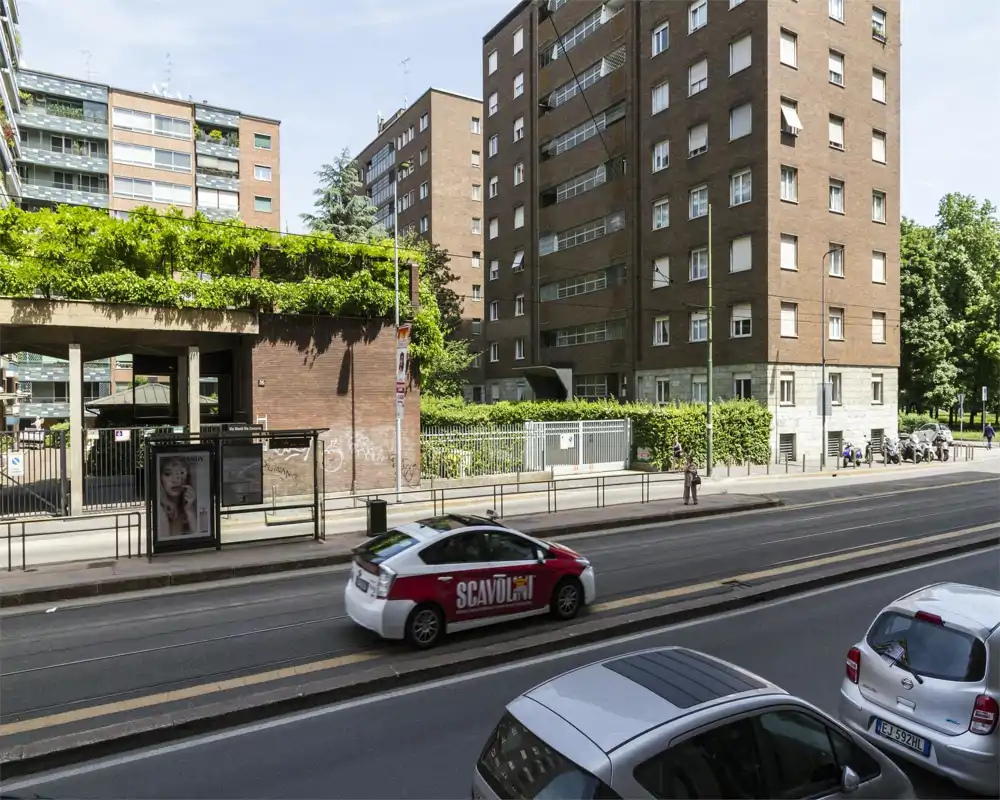 Vista esterna su strada con fermata del tram/autobus, auto parcheggiate e palazzi residenziali di più piani, con aree verdi lungo il marciapiede.