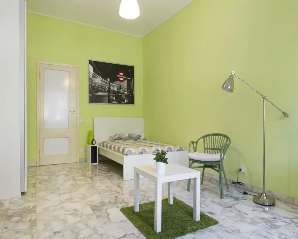 Camera da letto luminosa e moderna con letto piattaforma bianco, biancheria a fantasia e un piccolo tavolino su un tappeto verde. Parete verde chiaro, pavimento effetto marmo e lampada da lettura accanto a una sedia in vimini.