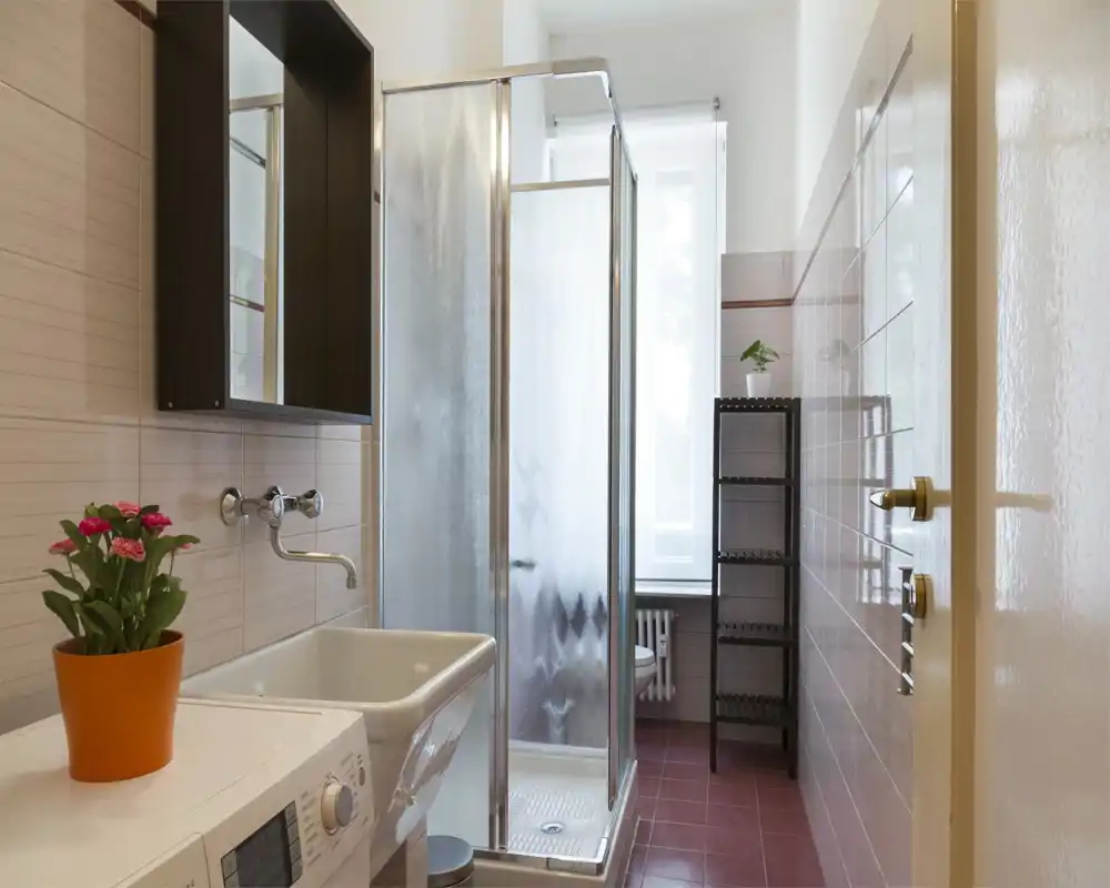 Baño moderno y luminoso con cabina de ducha, lavabo, lavadora y estantería; paredes alicatadas y una ventana que aporta luz natural.