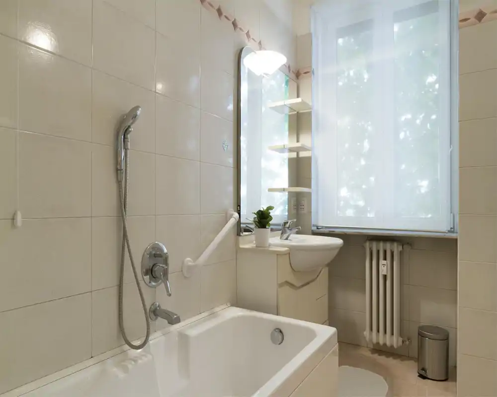 Baño limpio y luminoso con bañera, ducha de mano, lavabo y una ventana que aporta luz natural suave.