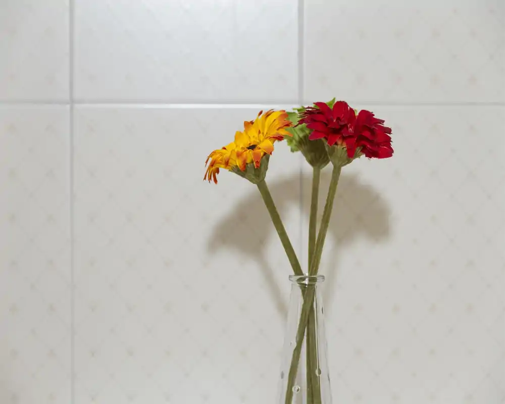 Plano detalle de tres flores decorativas en un jarrón de vidrio con un fondo de azulejos. La imagen destaca la composición y la textura de las flores, adecuada como detalle decorativo.