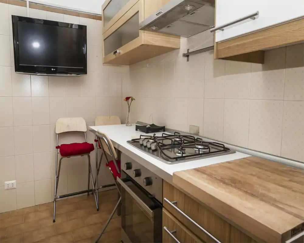 Cocina moderna y compacta con placa de gas, horno integrado y muebles de madera; pequeño mostrador con taburetes y TV en la pared.
