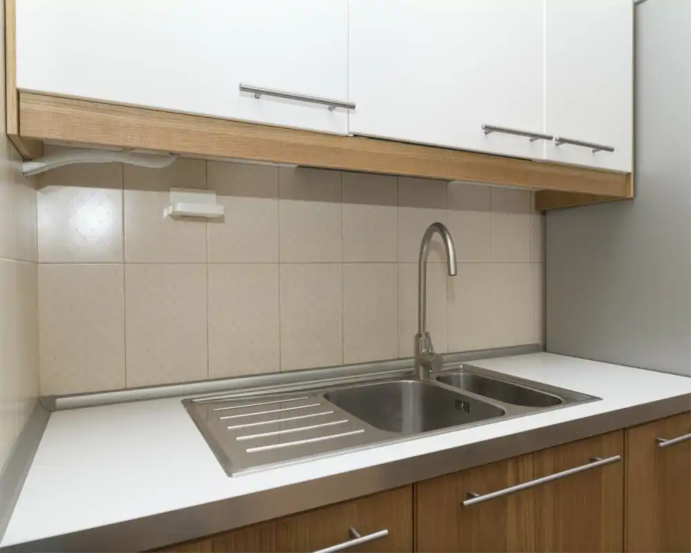 Área de cocina compacta y moderna con fregadero doble de acero inoxidable, encimera blanca minimalista y armarios de madera, iluminada de forma uniforme.