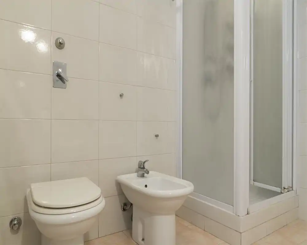 Baño limpio y funcional con inodoro, bidé y plato de ducha en esquina. Azulejos neutros y sanitarios sencillos que ofrecen un espacio práctico y de bajo mantenimiento.