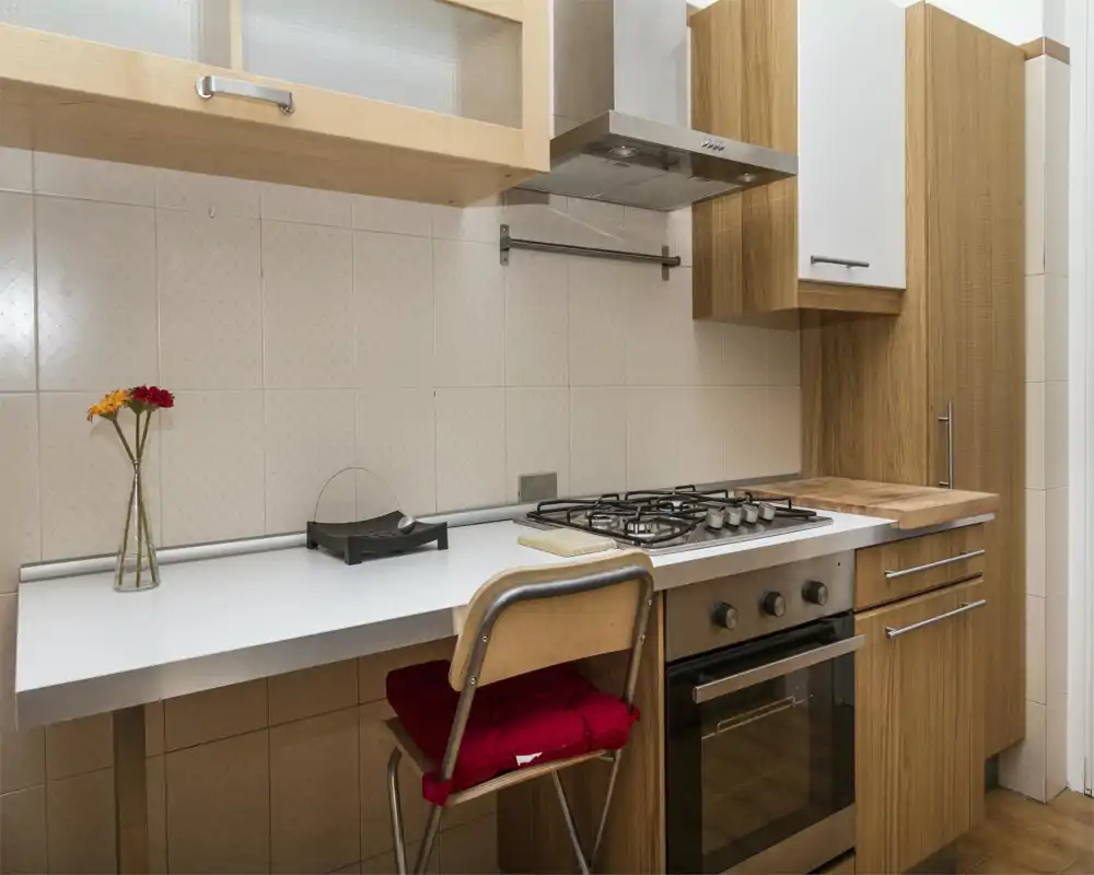 Cocina compacta y moderna con horno empotrado y placa de gas, muebles efecto madera y una pequeña encimera con taburete y jarrón decorativo.