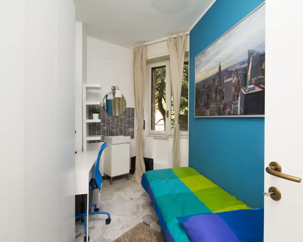 Small, well-lit bedroom with a single bed dressed in green and blue linens, a desk and chair beside a window, and contemporary wall art on a turquoise accent wall.