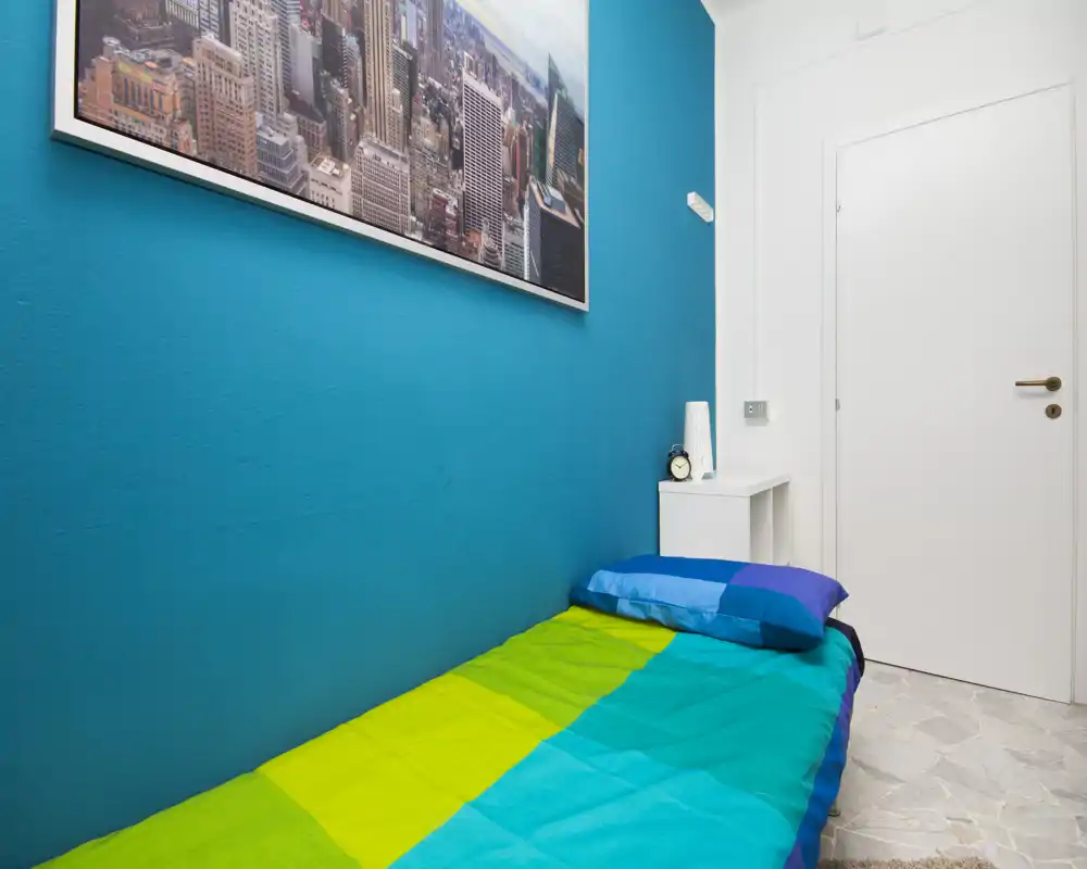 Compact single bedroom with a vibrant teal accent wall, colorful bedding and a small bedside table. Bright, tidy and suitable for a single guest or student.