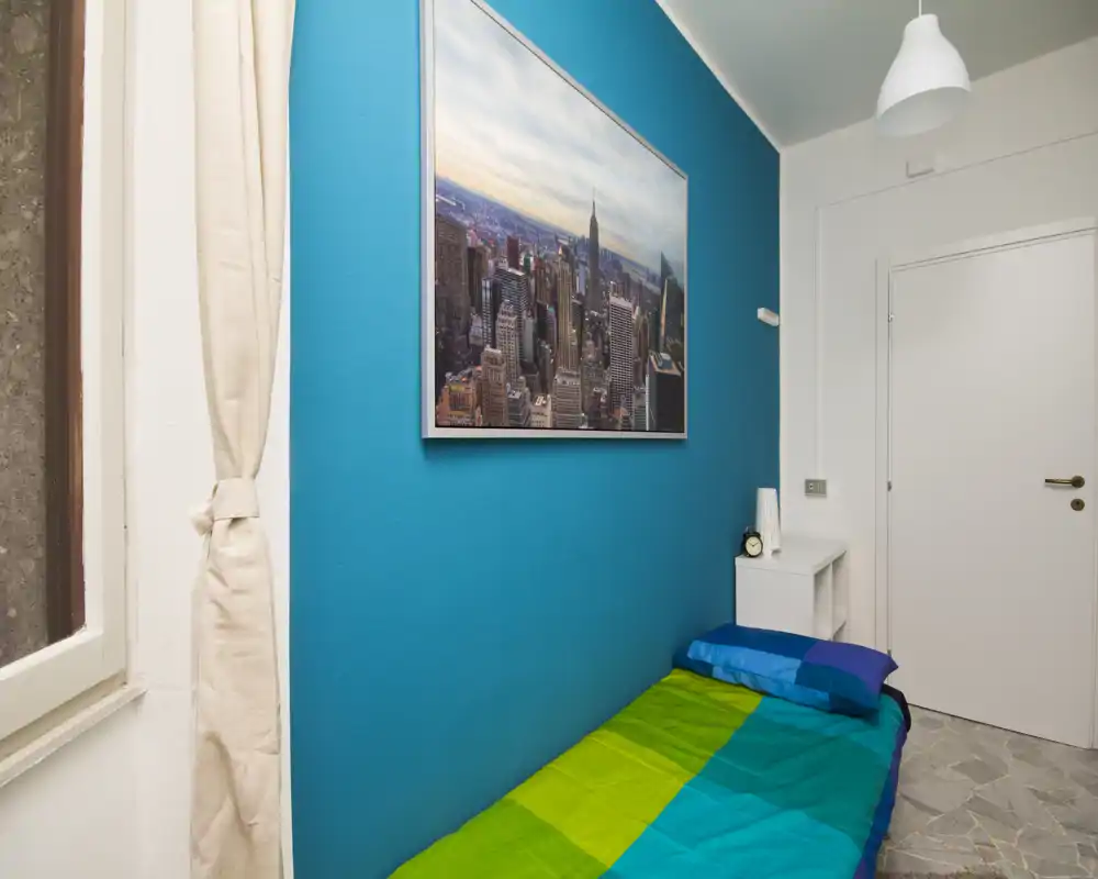 Compact single bedroom with a bright teal accent wall, colorful bedding and a small bedside table; tidy and well lit, suitable for a short-stay listing.