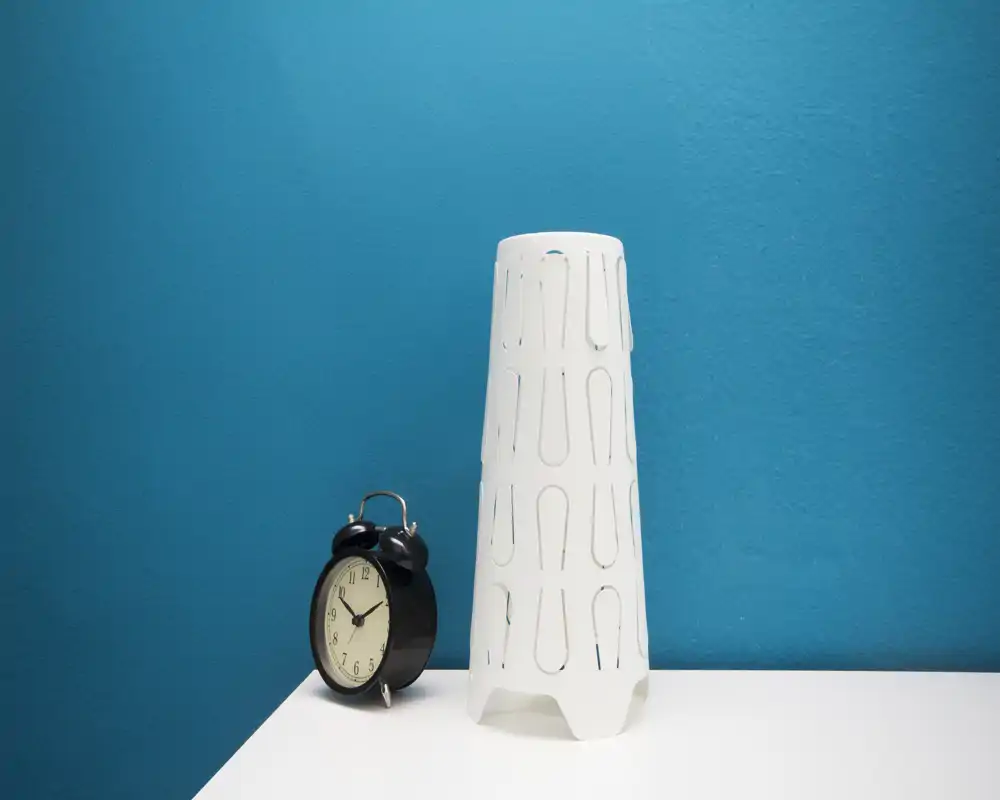Close-up still life on a bedside surface: a vintage-style black alarm clock and a tall white decorative lamp/cover against a vibrant blue wall. Good for highlighting small decor details and color contrast.