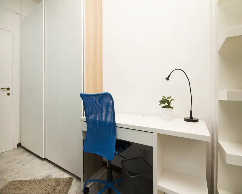Compact workspace corner with a white desk, blue ergonomic chair, desk lamp and a small potted plant. Bright, minimal and suitable to describe a study or home-office nook.