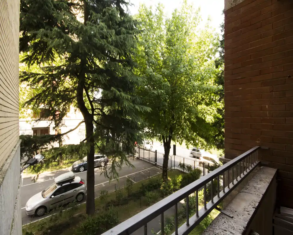 Sunny balcony view overlooking a tree-lined street with parked cars and a metal railing — ideal to show outdoor space and street outlook.
