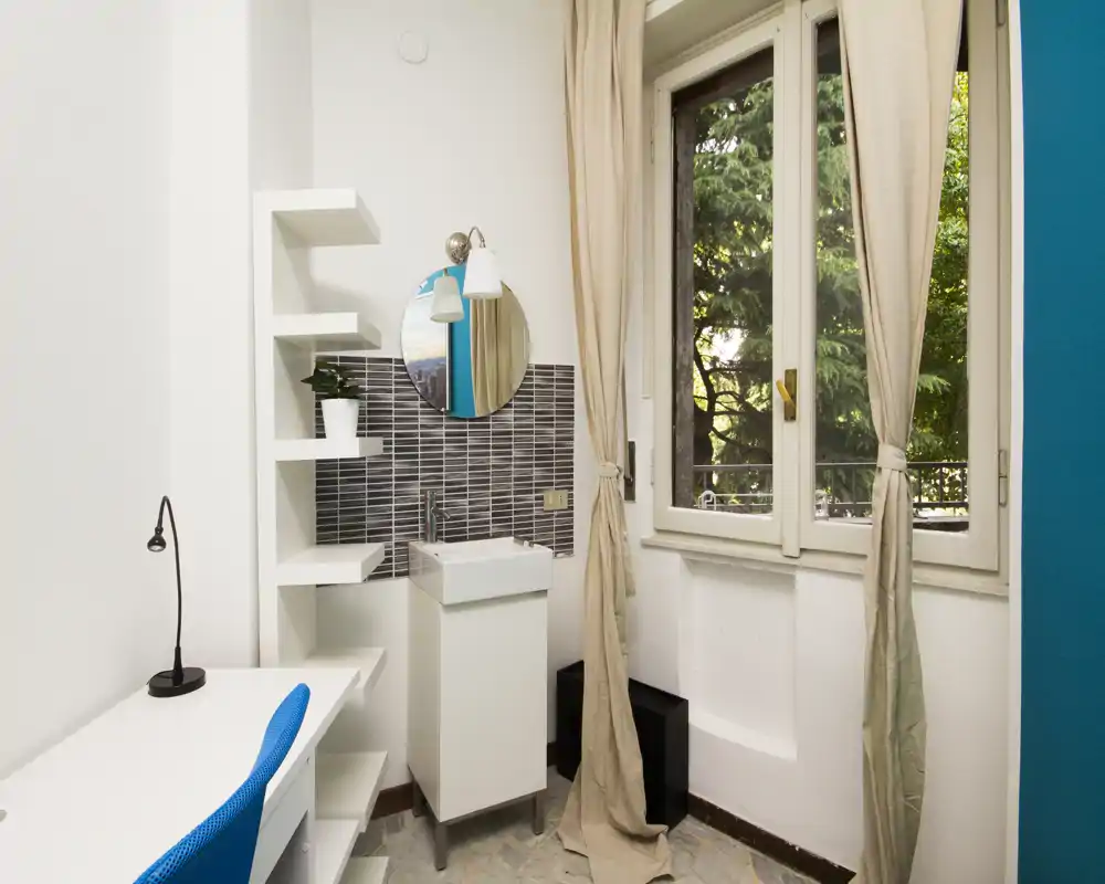Small bathroom corner with a compact sink, mirrored round vanity and tiled splashback beside a window letting in natural light.