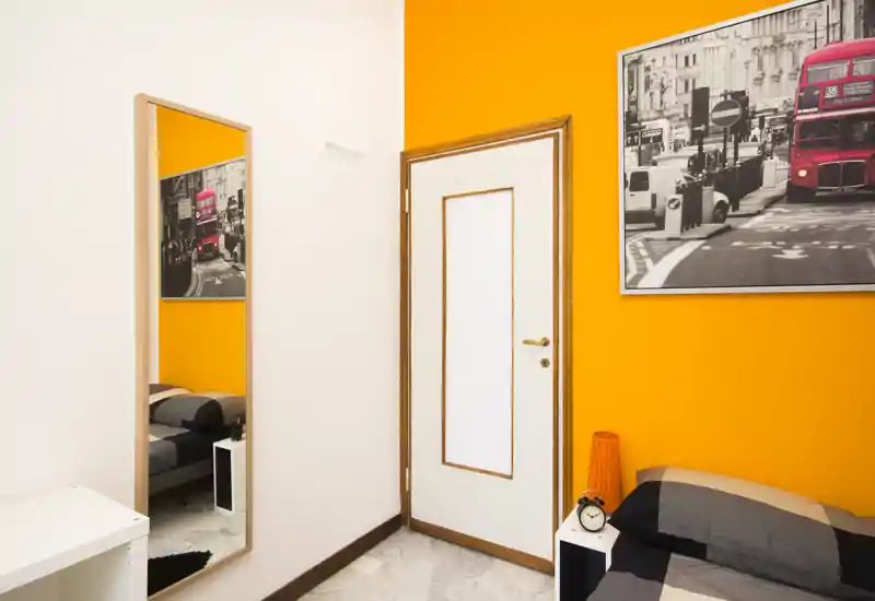 Dormitorio moderno y luminoso con cama individual bien hecha, mesita de noche y cuadro temático de Londres en una pared de acento naranja.