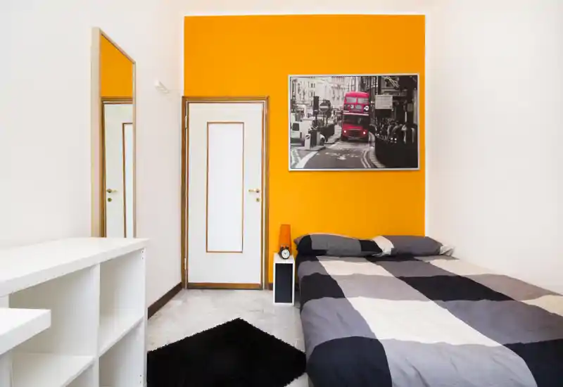 Dormitorio moderno y luminoso con pared naranja como acento, cama doble con ropa de cama estampada, mesita con lámpara y espejo de cuerpo entero — limpio, bien iluminado y atractivo para alquileres.