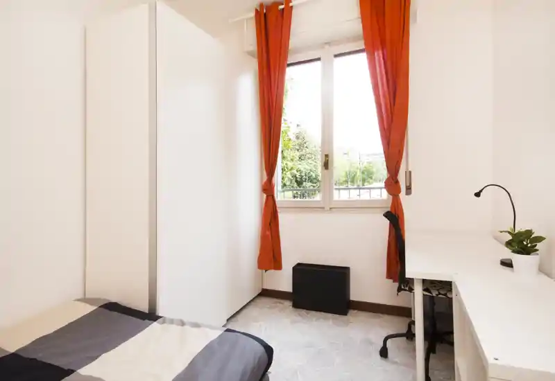 Dormitorio compacto y luminoso con cama individual, armario alto blanco y escritorio junto a la ventana. La luz natural y las cortinas naranjas crean un ambiente alegre, perfecto para estudiante o una persona sola.