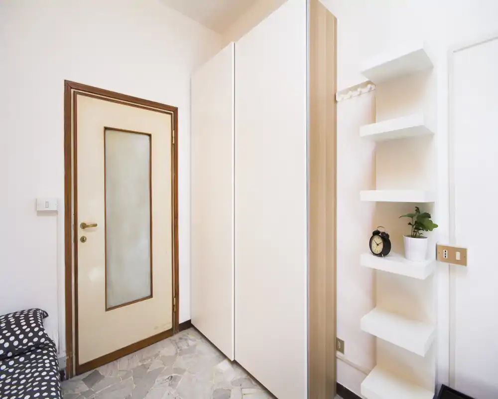 Coin de pièce compact avec un lit partiellement visible à gauche, une grande armoire et des étagères murales près de la porte. Lumineux, décor minimal et sol carrelé.