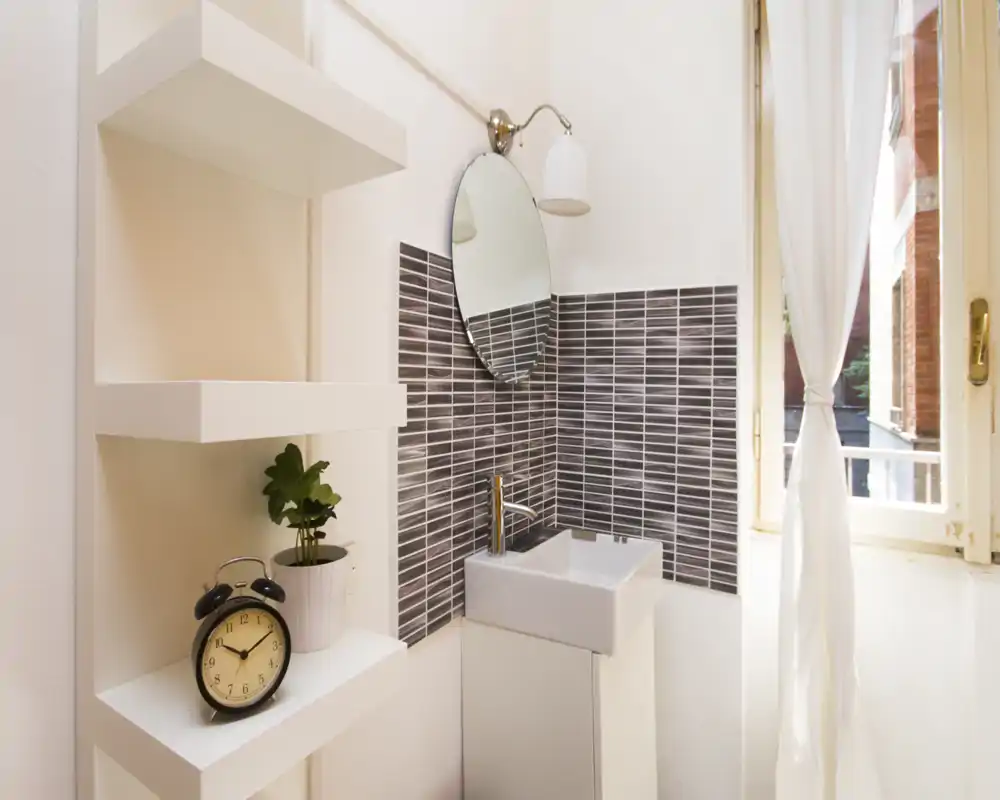Coin de salle de bains compact avec un petit lavabo, robinet moderne et miroir rond ; étagères blanches et une fenêtre apportant la lumière naturelle.