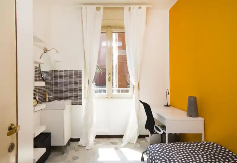 Chambre simple lumineuse avec bureau près de la fenêtre, rideaux blancs et un mur d'accent jaune; lit compact à motif à pois et lumière naturelle pour une ambiance aérée.