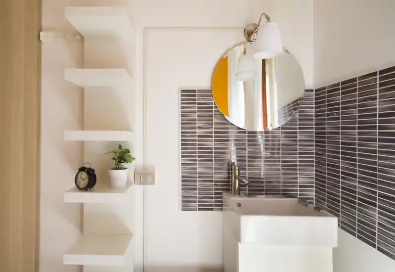 Small modern bathroom corner with a rectangular sink, glossy horizontal tile backsplash, round mirror and wall light; clean, bright and minimal.