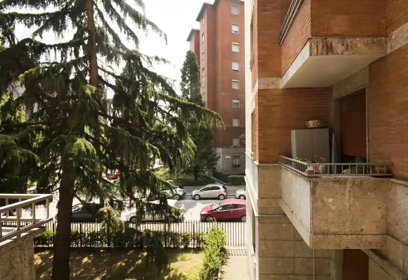 Sunny balcony view overlooking a tree-lined street with parked cars and neighboring brick buildings; small planter and railing visible, ideal to showcase an apartment’s outdoor space.