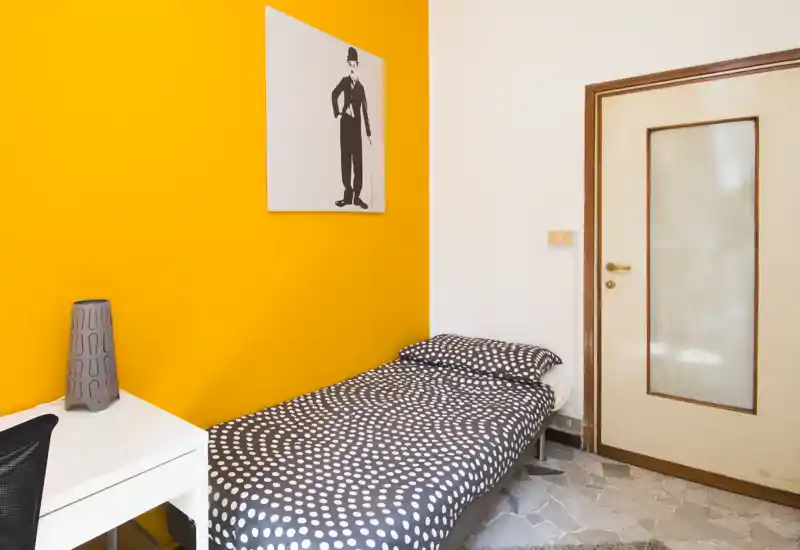 Chambre simple compacte avec lit à pois, mur jaune vif et petit bureau — propre, bien éclairée et idéale pour un étudiant ou une personne seule.