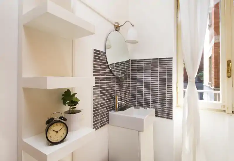 Compact bathroom corner with a small sink, modern faucet and round mirror; white shelving and a window provide natural light.