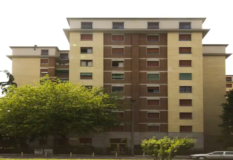 Exterior view of a multi-story residential building with a symmetrical brick and plaster façade, rows of closed shutters, and street-level entrance framed by trees.