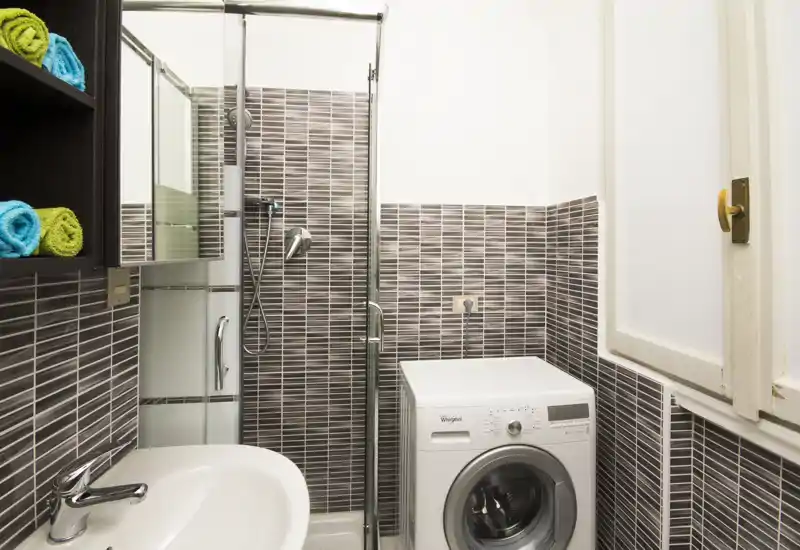 Compact modern bathroom with tiled walls, a glass shower enclosure, sink and a front-loading washing machine — bright and neatly arranged.