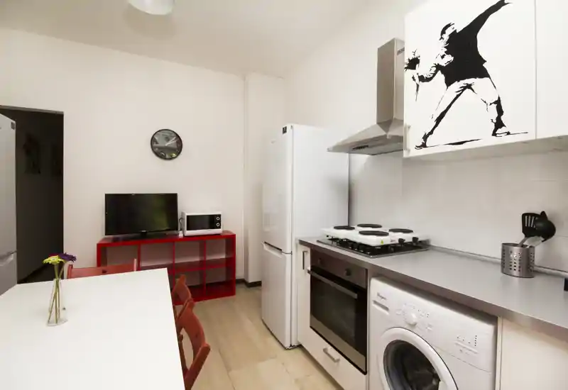 Compact modern kitchen with integrated appliances, a white countertop, washing machine under the counter, fridge and gas hob; dining table with red chairs and small decorative touches.