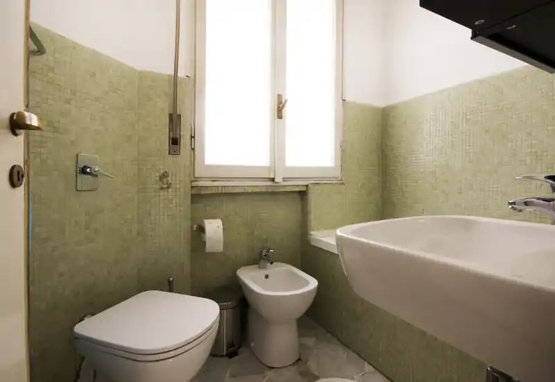 Compact bathroom with toilet, bidet and wall-mounted sink set against pale green mosaic tiles and a frosted window providing natural light.