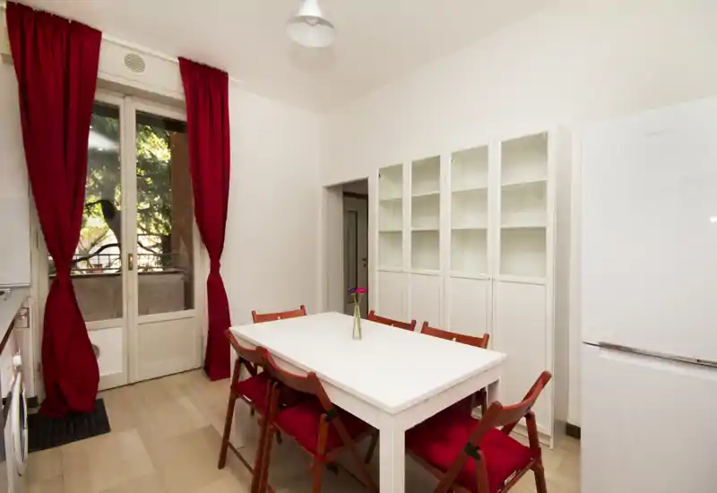 Bright, well-styled dining area with a white table, red-cushioned folding chairs and tall windows with red curtains leading to a small balcony.