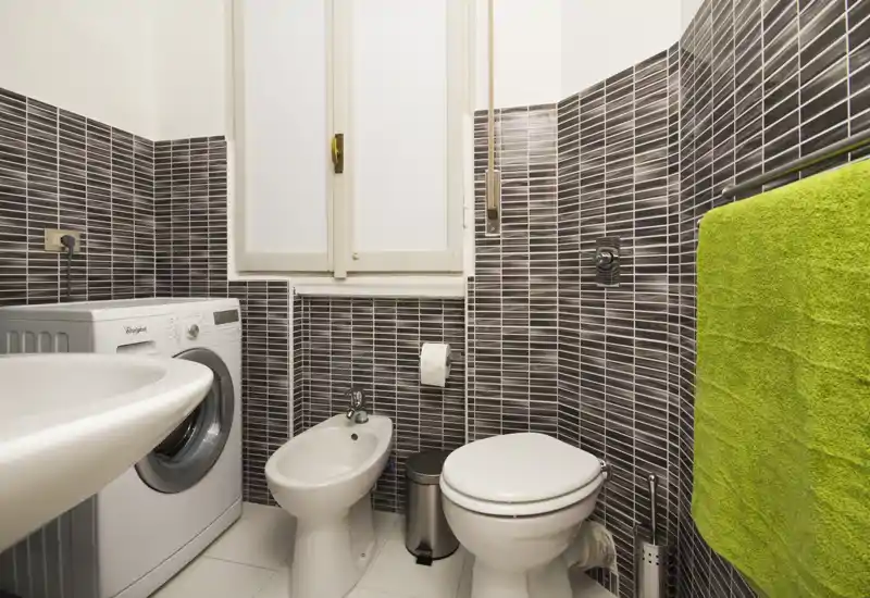Compact, modern bathroom with toilet, bidet, sink and a washing machine; dark horizontal mosaic tiles and a bright green towel add contrast.