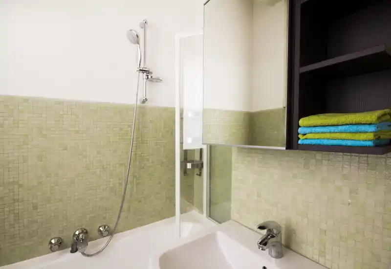Clean, modern bathroom corner with a bathtub, handheld shower, sink and mirrored cabinet against pale green mosaic tiles.