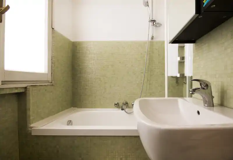 Bagno luminoso e pulito con vasca, doccia a parete e lavabo moderno con rubinetteria cromata su piastrelle a mosaico verde chiaro.