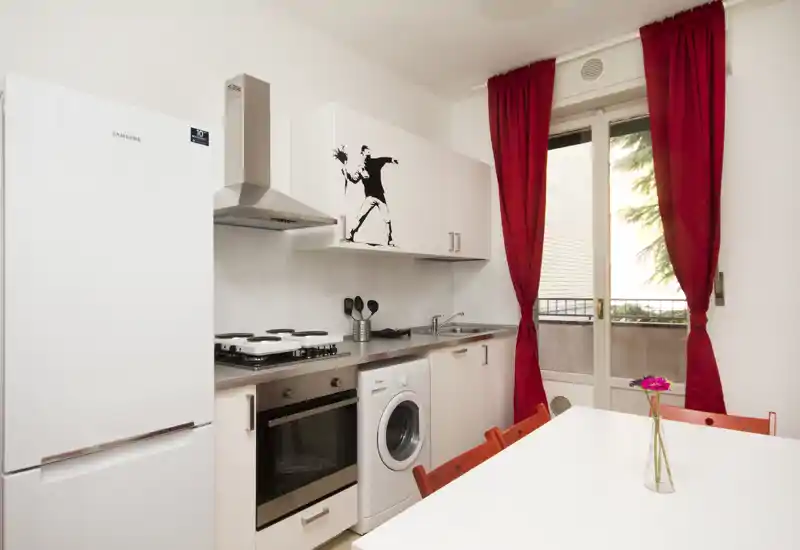 Compact modern kitchen with white cabinetry, built-in oven, stovetop, fridge and a washing machine; bright space with red curtains and a small dining table with a single flower.