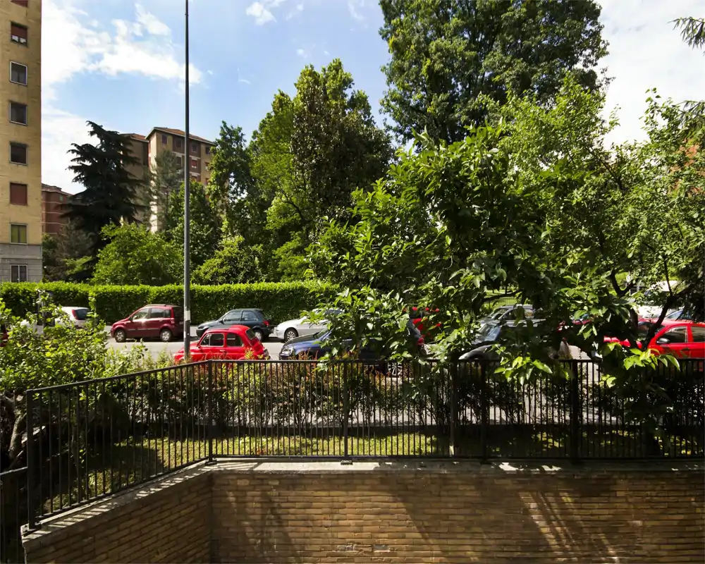 Sunny outdoor view from a lower-level terrace or garden: metal railing, trees and a street with parked cars beyond. Bright daytime scene with greenery and urban surroundings.