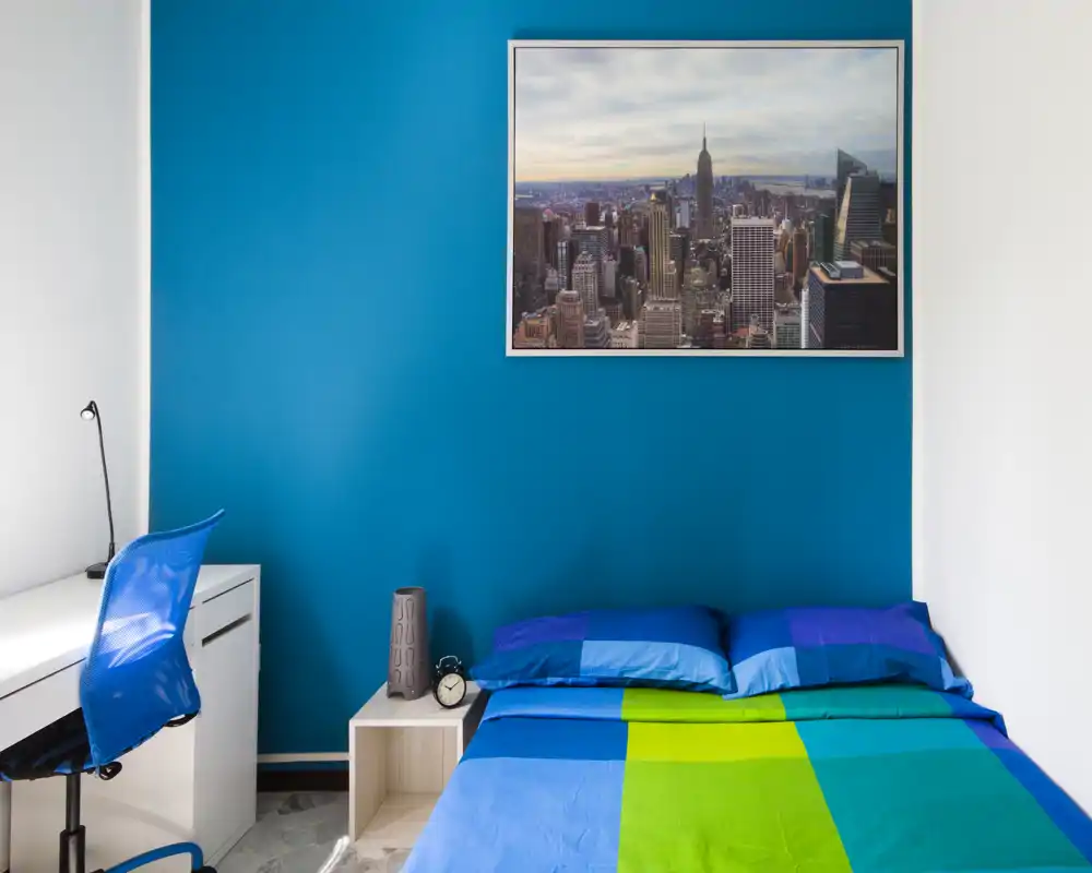 Bright, modern bedroom with a colorful double bed against a vivid blue accent wall, bedside table and a small work desk with chair — ideal for a single occupant or couple seeking a vibrant, compact room.