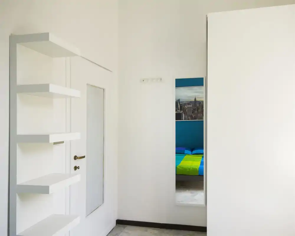 Bright, minimal entrance area with wall-mounted shelves and a full-length mirror reflecting a bedroom; clean white walls and tiled floor create a fresh, modern first impression.