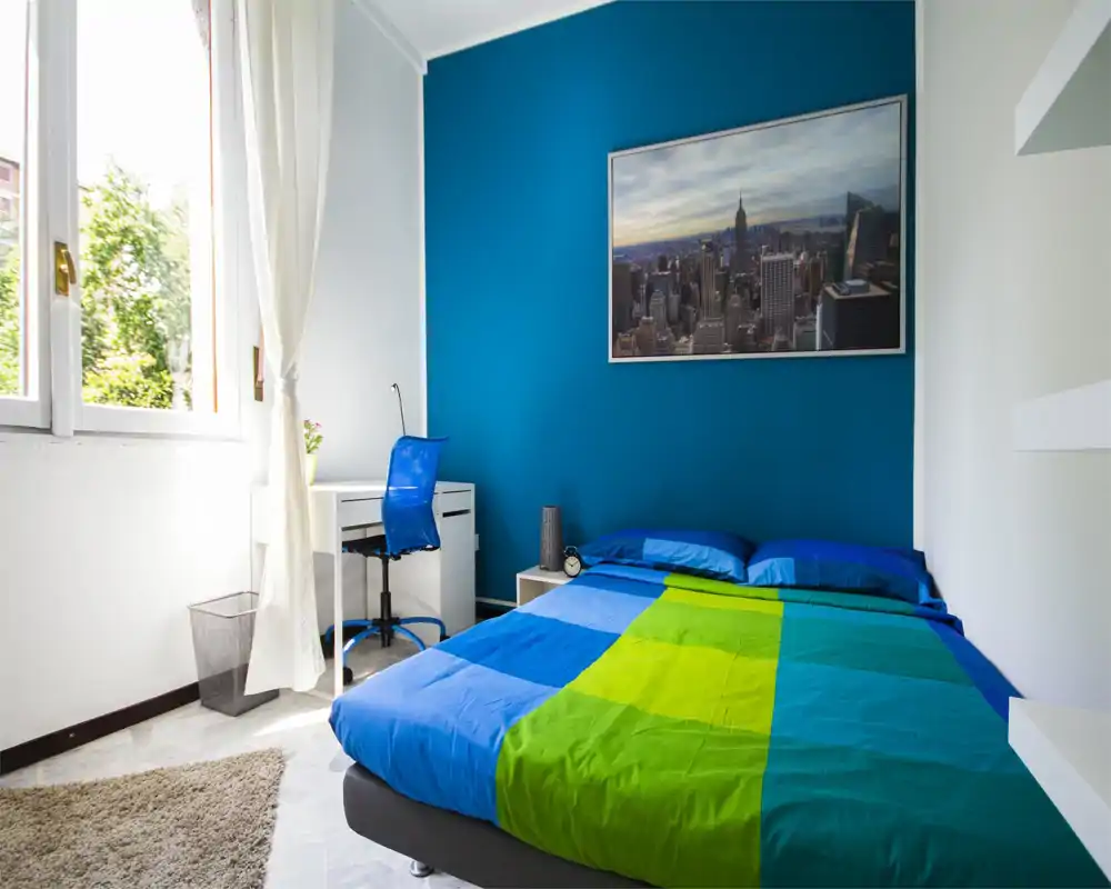 Bright, modern bedroom with a blue accent wall, double bed dressed in colorful bedding, a small desk and chair by the window and a large framed cityscape above the bed.