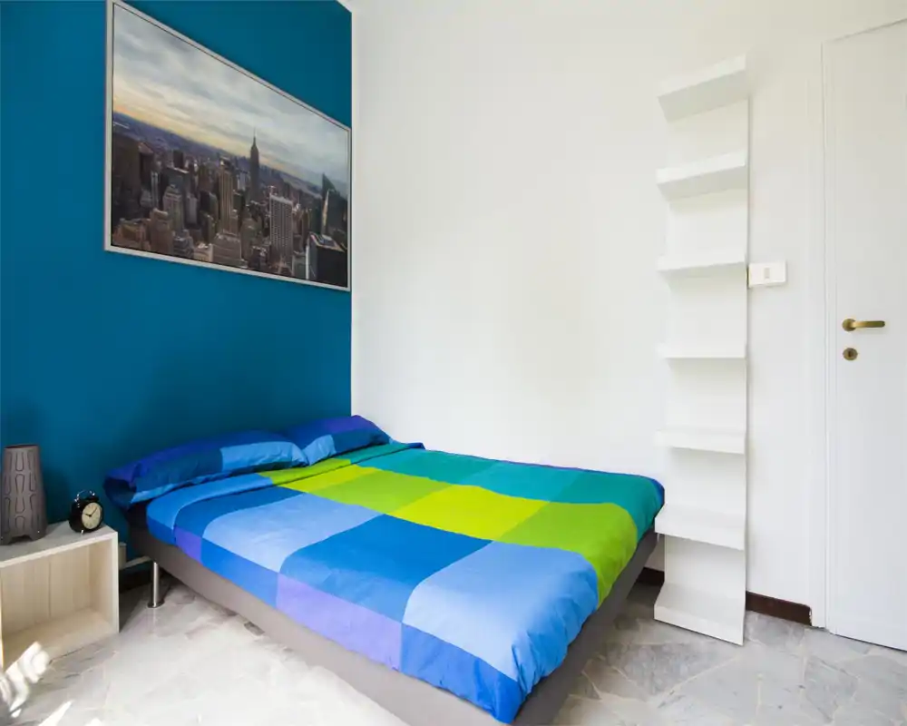 Bright, modern bedroom with a large bed dressed in colorful blue and green linens, a teal accent wall and a cityscape artwork above the headboard. Simple bedside table and shelving keep the space uncluttered and fresh.