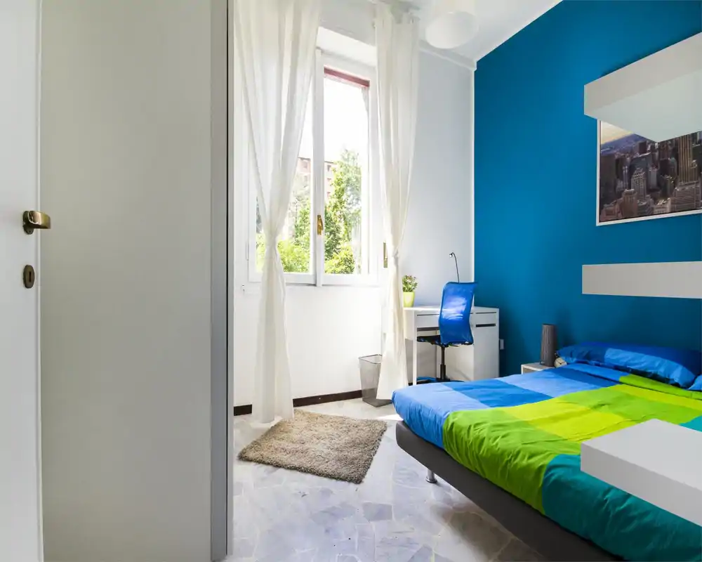 Bright, modern bedroom with a blue accent wall, a comfortable double bed with colorful bedding, and a small desk by the window — ideal for working or relaxing in natural light.