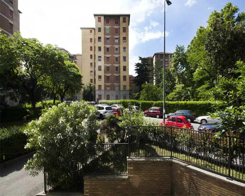 Bright exterior view of a residential courtyard with parking spaces, greenery and a mid-rise apartment building in the background.