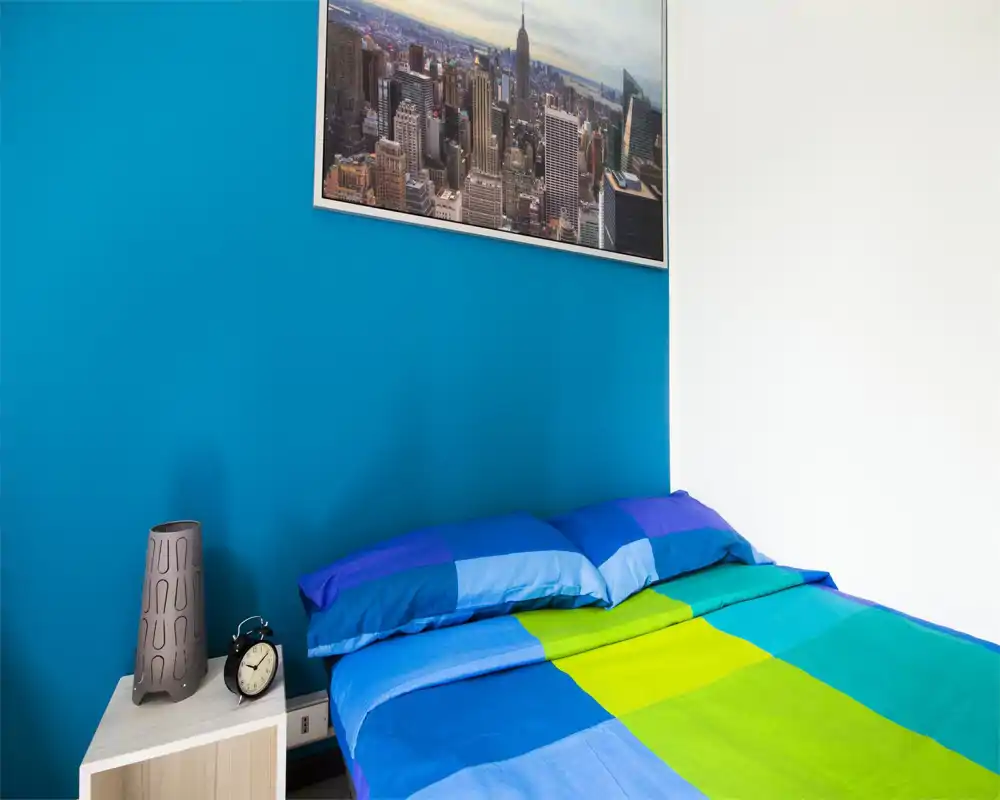 Bright, modern bedroom with a blue accent wall and a colorful bedspread; simple nightstand with lamp and alarm clock adds functional charm.